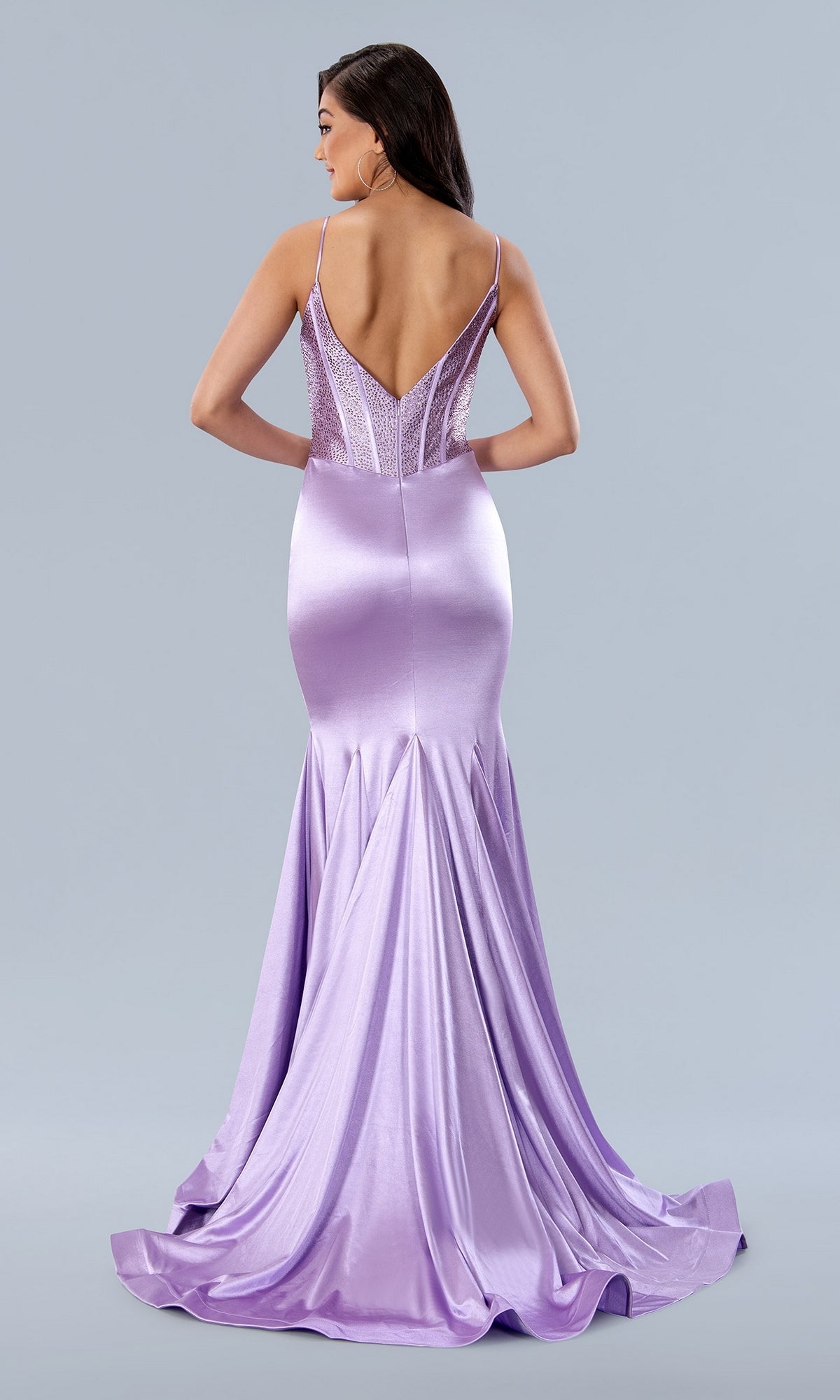 A woman stands against a light gray background wearing the Stella Couture 24198 Deep V-Back Long Prom Dress—a floor-length, lavender satin gown with thin straps, a beaded corset bodice, and a low V-shaped back.